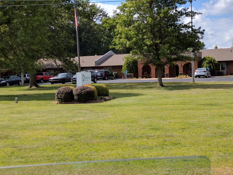 Exterior view of Fulton Nursing and Rehabilitation front entrance with lawn, trees, sign, and parked cars.