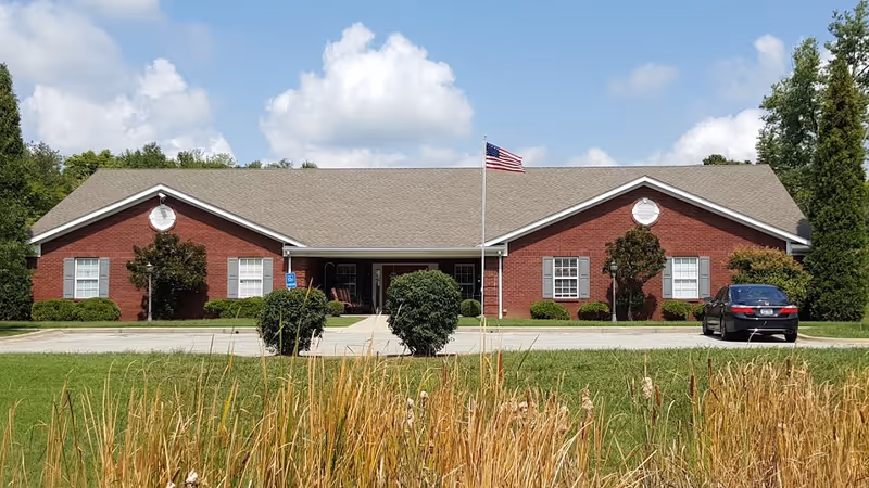 Front exterior view of a single-story red brick building with a gray shingled roof, white window shutters, and an American flag on a flagpole in front. There are bushes and trees around the building, a car parked on the right side, and a grassy area with tall dry plants in the foreground under a partly cloudy sky.