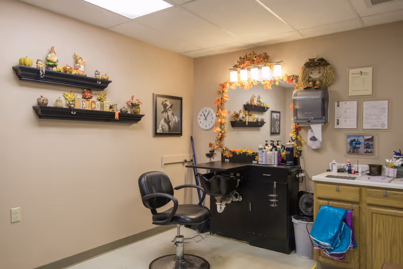 A small salon/styling station inside a senior facility with a barber chair, wash sink, mirror decorated with autumn garlands, and shelves of seasonal decor.