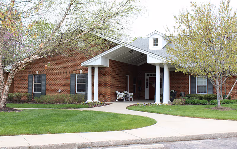 Brick senior living building front with a covered entrance supported by white columns, a curved walkway, and landscaped lawn and trees.