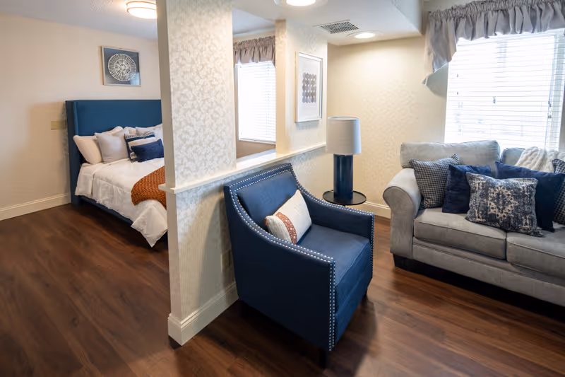 A cozy senior living space with a blue armchair and a gray sofa adorned with multiple decorative pillows. The room features wood flooring, a window with blinds and a valance, a side table with a lamp, and a partial view of a bedroom with a blue upholstered headboard and neatly made bed with white and orange bedding.