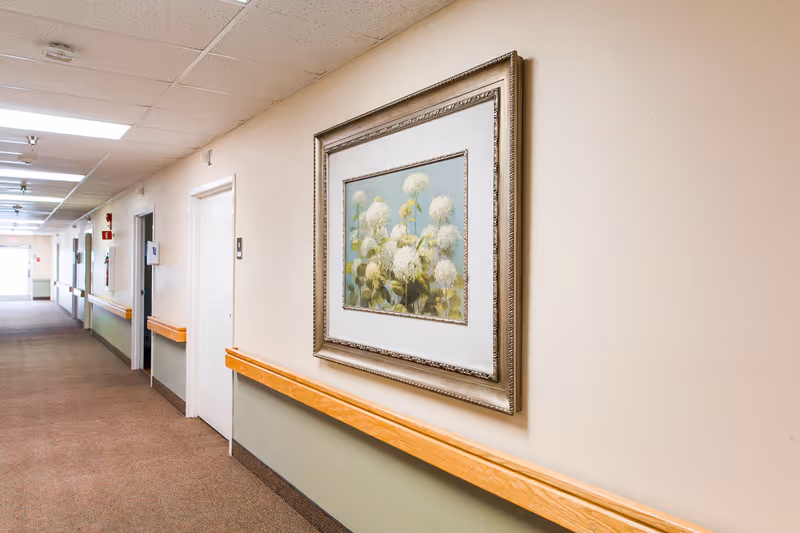A long indoor hallway with beige walls and carpeted floor. The hallway has wooden handrails on both sides and several doors along the left wall. A framed painting of white flowers with a light blue background is hung on the right wall. The ceiling has fluorescent lighting panels.