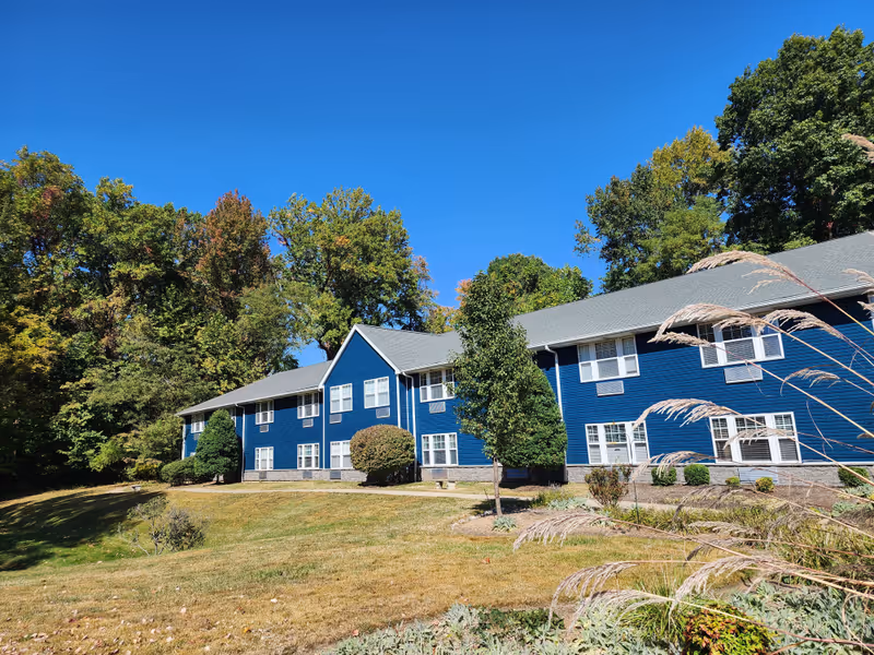 Exterior view of a two-story blue building with multiple windows, surrounded by trees and a grassy lawn under a clear blue sky.