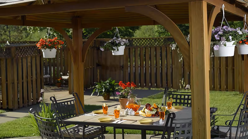 Outdoor wooden gazebo with a dining table set for four, featuring glasses of iced tea with lemon, a pitcher of iced tea, a fruit platter, and a flower pot centerpiece. Hanging flower pots with colorful blooms are suspended from the gazebo roof, and a wooden fence and green lawn are visible in the background.