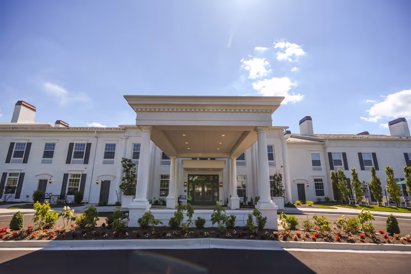 Front exterior view of a large white senior living facility building with a covered entrance supported by columns, landscaped garden beds with flowers and shrubs, and a clear blue sky with some clouds.