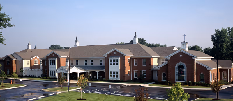 Exterior view of Franciscan Health Care Center, a large brick building with multiple peaked roofs and white trim, surrounded by a parking lot and landscaped greenery under a clear sky.