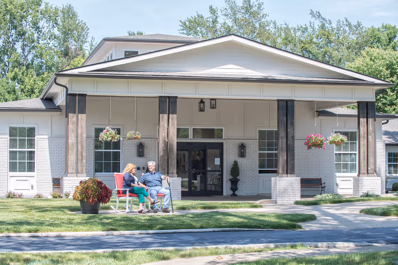 Two seniors sit in red chairs outside a senior living facility entrance.
