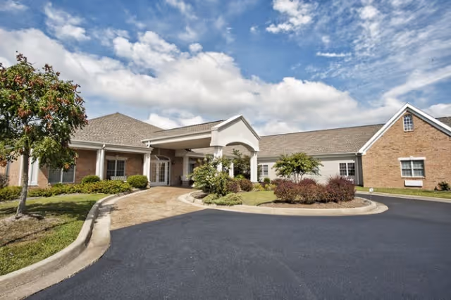Front exterior view of a single-story brick building with a covered entrance, surrounded by landscaped greenery and a curved driveway under a partly cloudy sky.