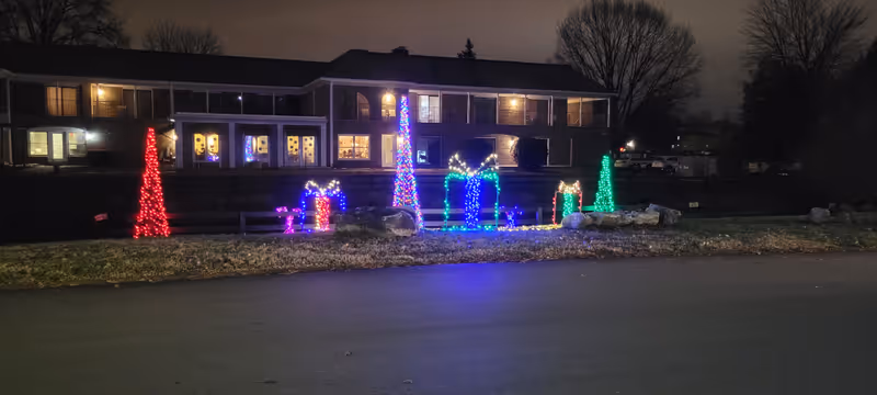 Night view of a two-story brick assisted living building front with colorful holiday lights and illuminated gift and tree shapes on the lawn.