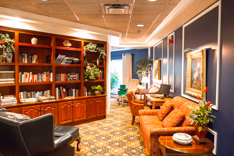 A cozy interior lounge area with a patterned carpet, a wooden bookshelf filled with books and decorative plants, a black leather armchair, a red patterned sofa with matching cushions, a wooden side table with a plant and decorative items, a desk with a computer, and framed artwork on dark blue walls.