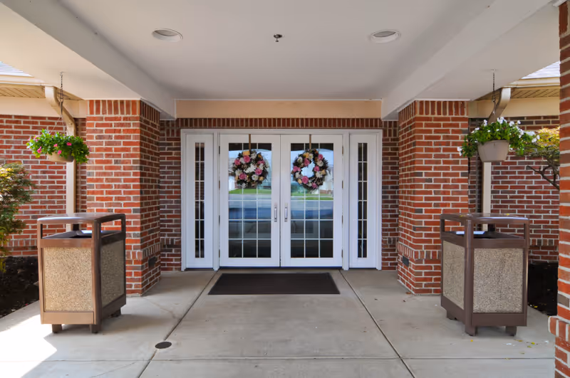 Entrance to a building with double glass doors decorated with floral wreaths. The entrance is framed by red brick pillars and has two hanging flower pots on either side. There are two trash bins positioned near the pillars on a concrete walkway.