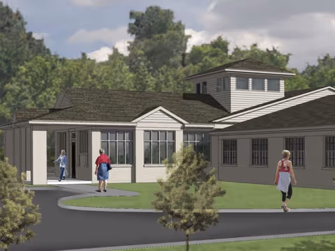 Exterior view of a single-story senior living facility building with a covered entrance and several windows. Three people are walking near the building on a paved path surrounded by grass and small trees. The sky is partly cloudy with trees in the background.