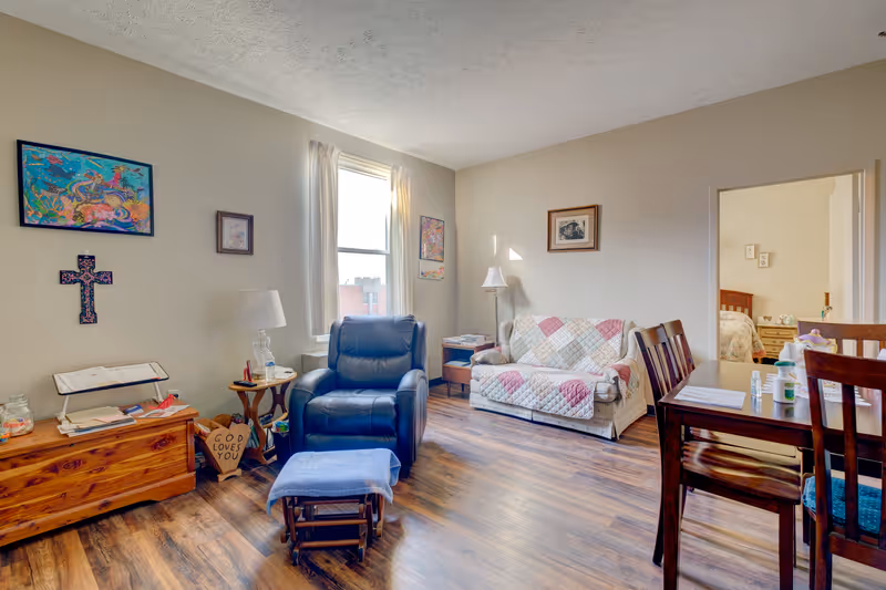 A cozy living room with a blue recliner chair and matching footrest, a wooden chest with a lamp and books, a small side table, a couch covered with a patchwork quilt, and a wooden dining table with chairs. The walls are decorated with colorful artwork and a cross. A doorway leads to a bedroom with a bed and nightstand visible.