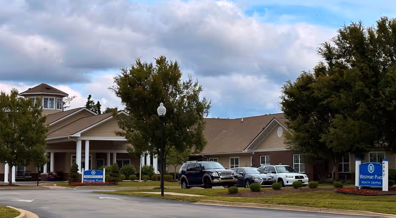 Exterior view of Westport Place Health Campus building with a driveway, several parked cars, trees, and a blue sign displaying the facility name. The sky is partly cloudy.