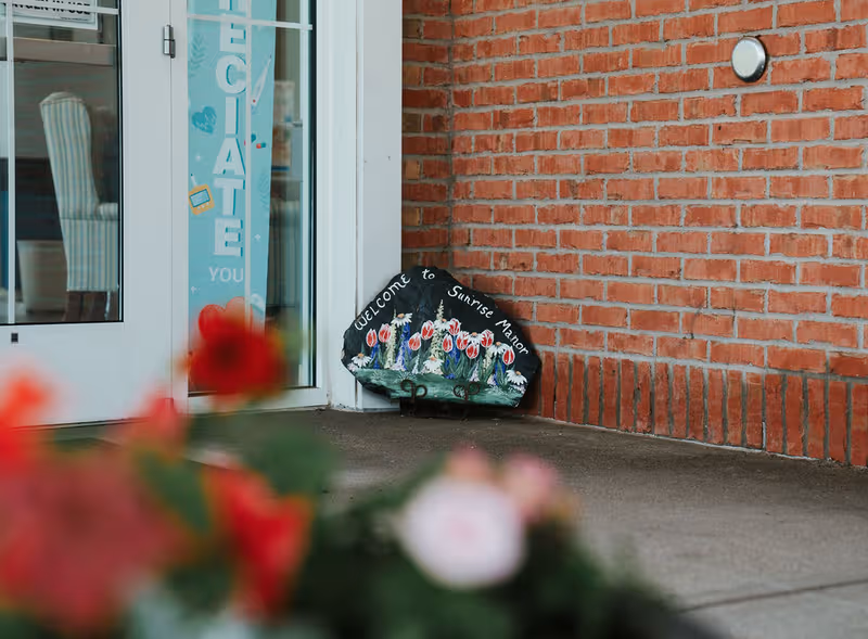 Entrance to Sunrise Manor Nursing Home with brick exterior, glass door, and a decorative sign.