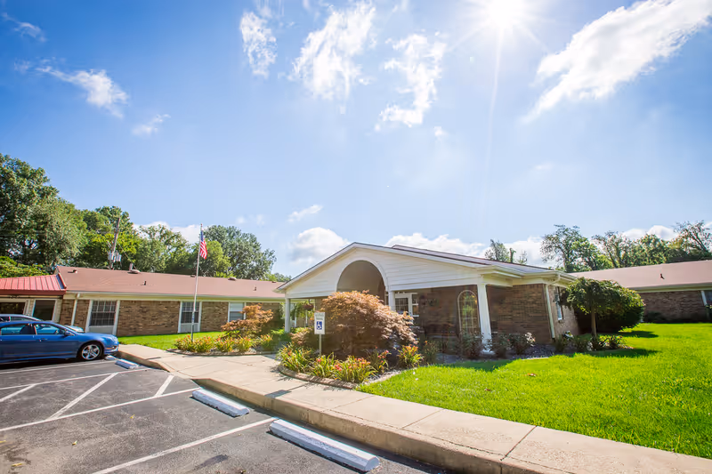 Exterior view of a single-story brick building with a covered entrance, surrounded by green grass and landscaping under a bright sunny sky. A blue car is parked in the parking lot near the building, and an American flag is visible on a flagpole.