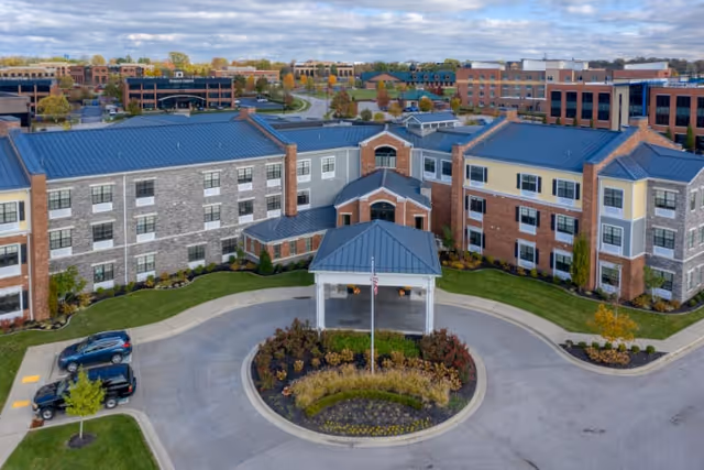 Aerial view of Cedarhurst of Beaumont senior living facility showing a large three-story building with a mix of brick and stone exterior, a covered entrance with a circular driveway, landscaped greenery, and parked cars. The background includes other buildings and a partly cloudy sky.