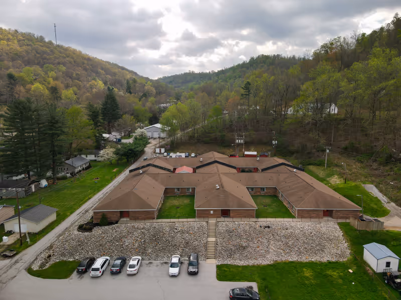 Aerial view of a single-story brick building with a brown roof, surrounded by green trees and hills under a cloudy sky. There are several cars parked in front of the building and a small road leading to it.