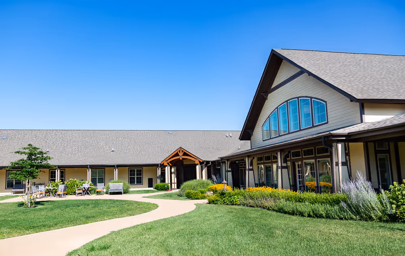 Exterior view of The Legacy at English Station facility showing a large building with multiple windows, a peaked roof, and a covered entrance. There is a well-maintained lawn with a curved concrete pathway, outdoor seating with chairs and tables, and landscaped flower beds under a clear blue sky.