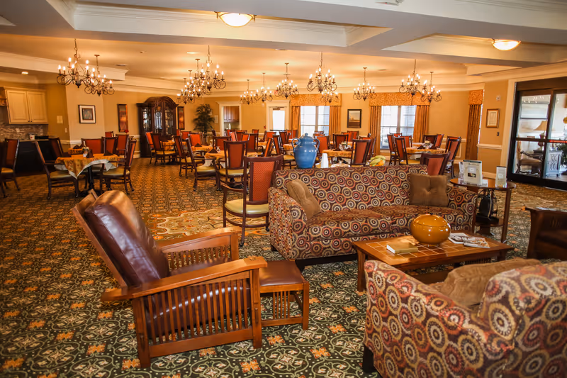 A spacious and warmly lit dining room with multiple tables and chairs arranged for meals. The room features patterned carpet, chandeliers hanging from the ceiling, and a sitting area with patterned sofas and wooden armchairs in the foreground. Large windows with curtains and a glass door leading outside are visible in the background.
