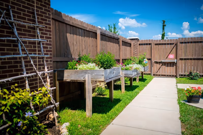 Outdoor garden area with raised wooden planters filled with green plants and flowers, a wooden fence surrounding the space, a brick wall on the left, a concrete pathway, and a clear blue sky with some clouds.