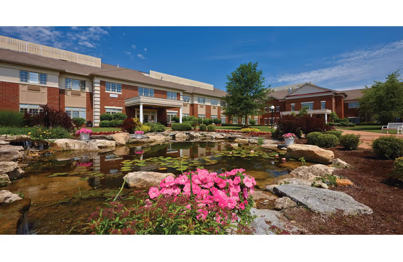 A landscaped outdoor area featuring a pond with rocks and flowers in front of a senior living facility.