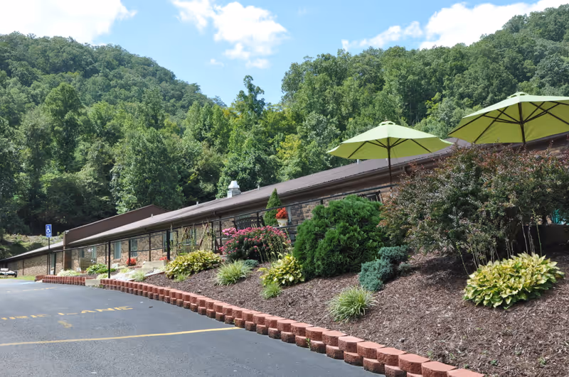 Exterior view of Good Shepherd Health and Rehabilitation Center showing a single-story brick building with a landscaped garden in front, including bushes, flowers, and two green patio umbrellas. The building is surrounded by lush green trees and hills under a partly cloudy sky. A paved parking area with a handicapped parking sign is visible in the foreground.