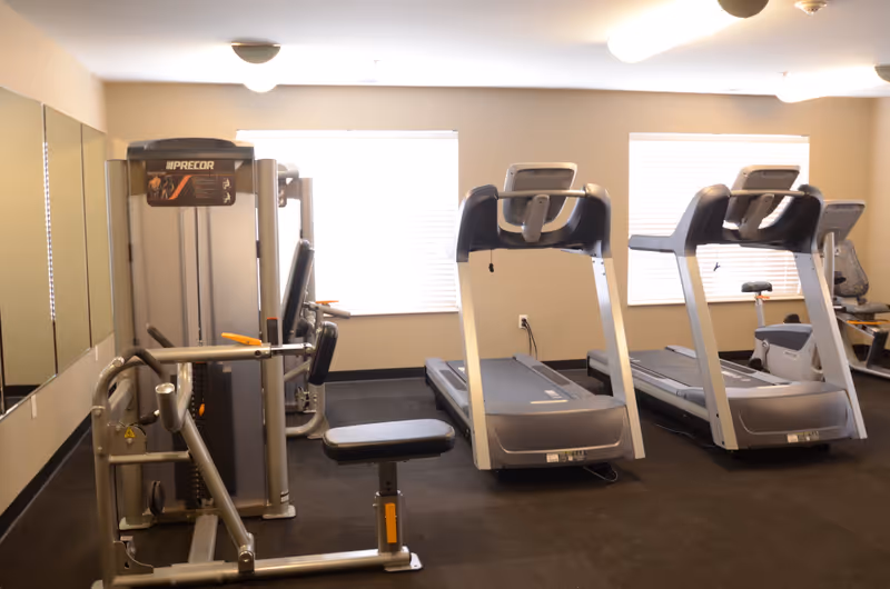 A fitness room with exercise equipment including two treadmills, a weight machine, and a stationary bike. The room has beige walls, two windows with blinds, and mirrors on one wall.