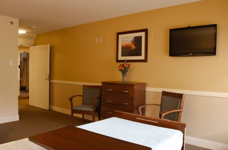 A senior living bedroom with a bed, wooden dresser, two chairs, wall-mounted TV and framed artwork on a beige wall.