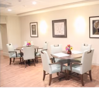 A bright dining area with light-colored walls and wooden flooring featuring two round tables, each surrounded by four cushioned chairs. The tables have small floral centerpieces, and framed artwork is displayed on the walls.