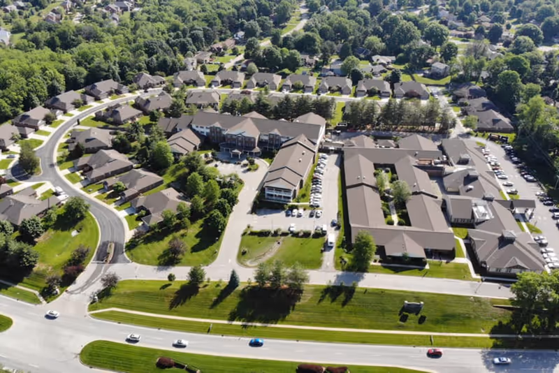 Aerial view of a memory care campus and surrounding residential neighborhood with multiple buildings, parking lots, and tree-lined streets.