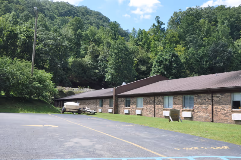 Single-story brick building with a brown metal roof, a parking lot and a trailer-mounted boat in front of a tree-covered hillside.