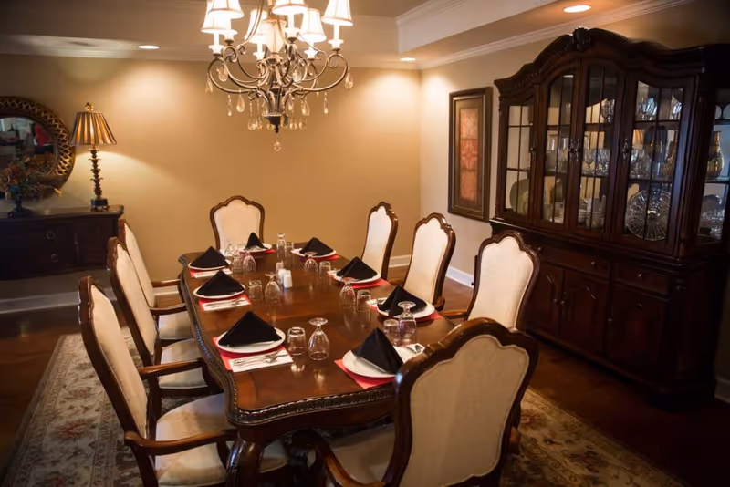 A formal dining room with a large wooden dining table set for eight people. Each place setting has a white plate, black folded napkin, glassware, and silverware. The room features upholstered dining chairs, a chandelier hanging above the table, a wooden china cabinet filled with glassware and dishes, a sideboard with a lamp and decorative mirror, and framed artwork on the wall.