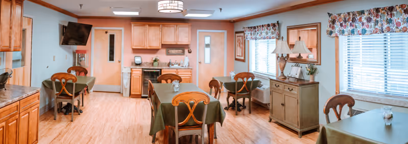 A dining room in Letcher Manor with several tables covered in green tablecloths, wooden chairs, a sideboard with two lamps and a mirror, windows with floral valances, and a kitchenette area with wooden cabinets and a wall-mounted TV.