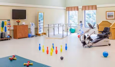 Bright therapy room with exercise equipment, parallel bars, a stationary bike, and bowling pins.
