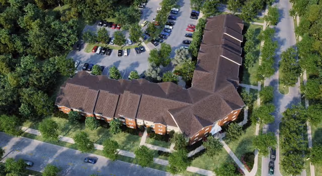 Aerial view of Hamburg Senior Residence, a large L-shaped brick building with a brown roof surrounded by trees and greenery. There are parking lots with cars around the building and sidewalks leading to the entrances.