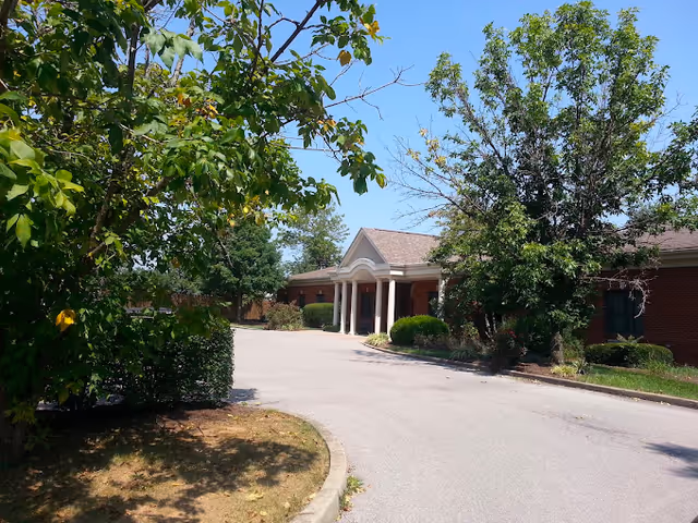 Exterior view of a single-story brick building with a covered entrance supported by white columns, surrounded by trees and bushes under a clear blue sky.