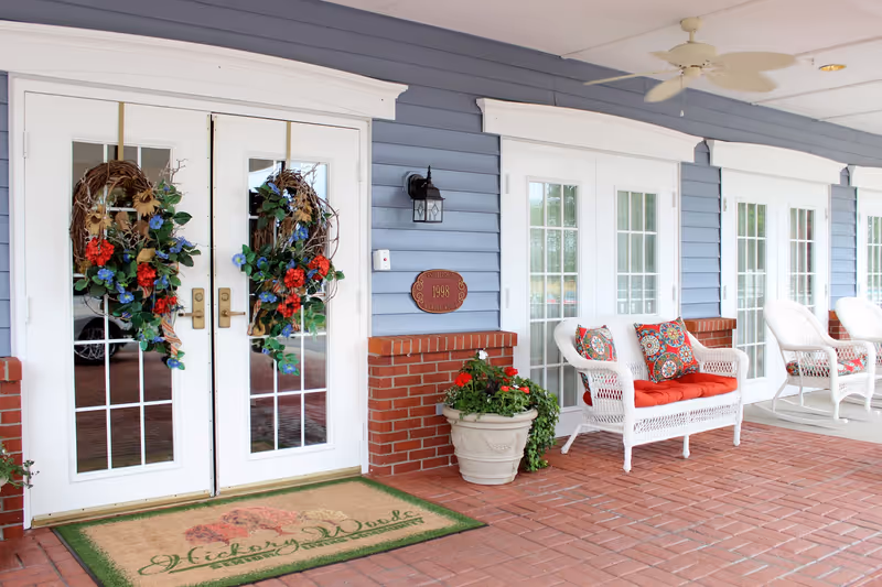 Entrance of Hickory Woods Senior Living Community featuring white double doors with floral wreaths, a welcome mat, brick flooring, blue siding, white wicker chairs with colorful cushions, and potted plants.