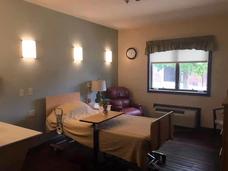 Hospital-style patient room with a single bed, chair, window, and bedside table.