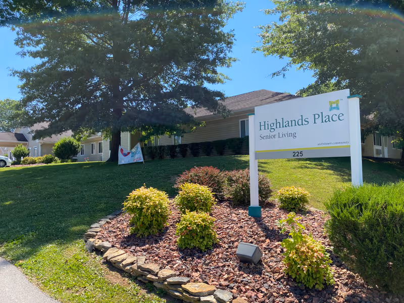 Exterior view of Highlands Place Senior Living with sign, trees, and landscaping.