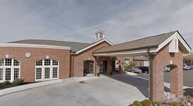 Exterior view of a brick building with a covered entrance and a circular driveway. Several cars are parked in the parking lot adjacent to the building under a partly cloudy sky.