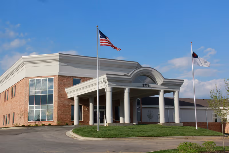 Entrance of a brick building with white columns and two flagpoles flying flags under a blue sky.