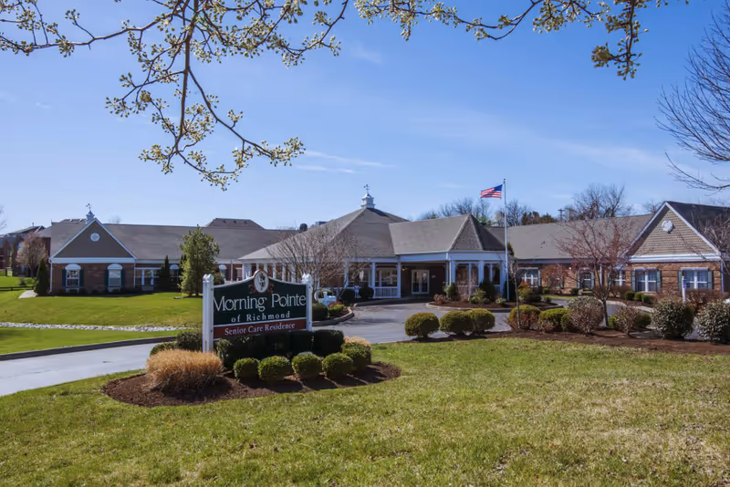 Exterior view of Morning Pointe of Richmond Senior Care Residence building with a well-maintained lawn, bushes, and an American flag on a flagpole. The building has a pitched roof and a covered entrance.
