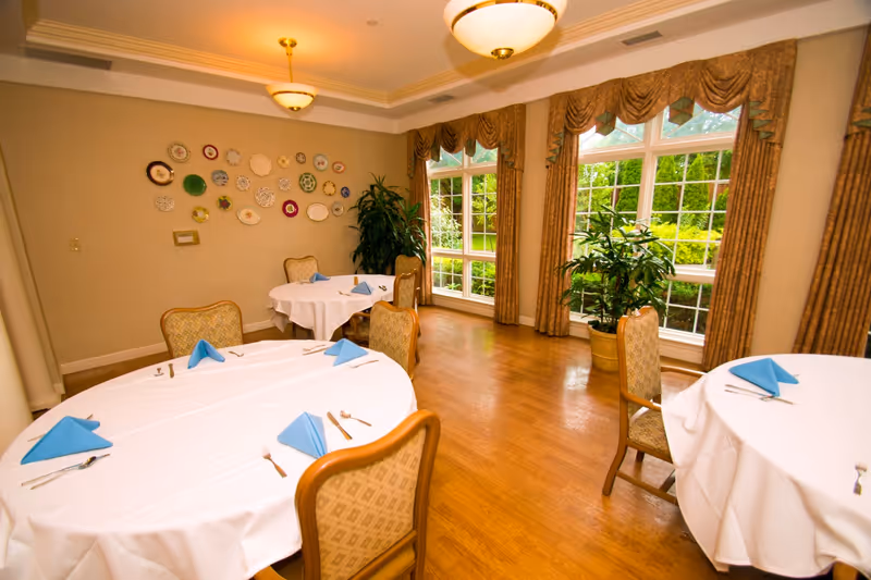 A dining room with round tables covered in white tablecloths, each set with blue folded napkins and silverware. The room has large windows with brown drapes, allowing natural light to brighten the space. Decorative plates are arranged on one wall, and there are potted plants near the windows. The floor is wooden, and ceiling lights provide additional illumination.