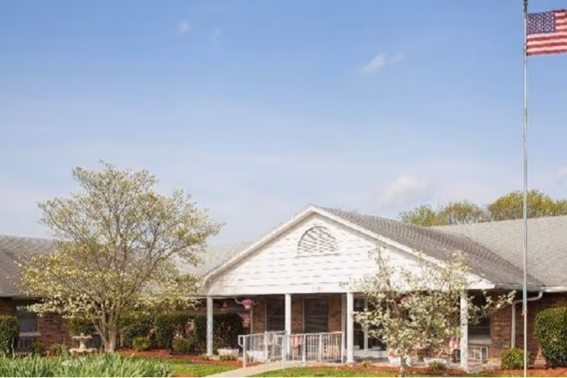 Exterior view of a single-story brick building with a white entrance porch, surrounded by trees and greenery under a clear blue sky. An American flag is flying on a flagpole to the right of the entrance.