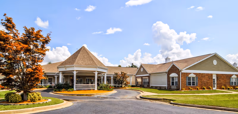 Front entrance of The Neighborhood at Paducah senior living facility showing a gazebo-style porte-cochere, circular driveway and landscaped grounds under a blue sky.