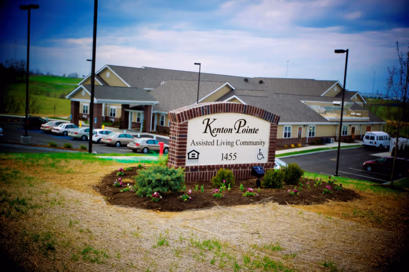 Brick sign reading "Kenton Pointe Assisted Living Community" sits in a landscaped area with the facility building and parked cars visible behind it.