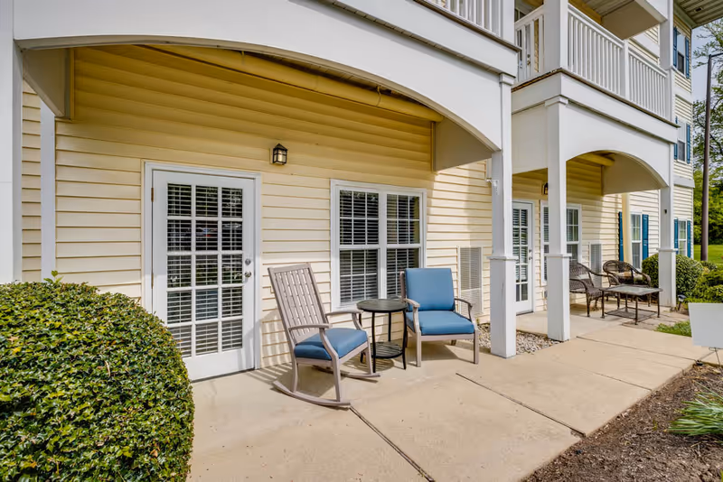 Outdoor patio area of a senior living facility with beige siding, white-framed glass doors and windows, blue cushioned chairs, a small round table, and neatly trimmed bushes along a concrete walkway.