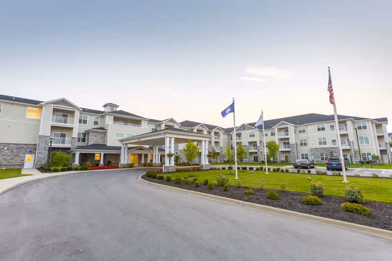 Exterior view of a large, modern senior living facility with three stories, multiple balconies, a covered entrance, landscaped grounds, and three flagpoles displaying flags in front of the building during early evening.
