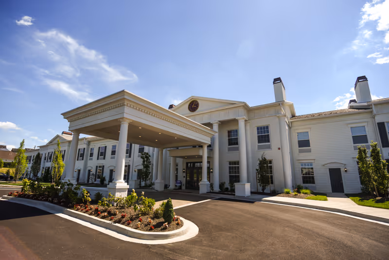 Exterior view of a large, elegant senior living facility building with white columns and a covered entrance driveway. The building has multiple windows and is surrounded by landscaped greenery and a clear blue sky.